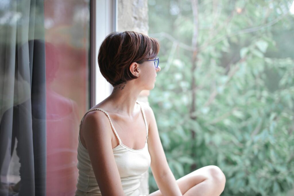 Side view of calm thoughtful young female in casual sleepwear sitting on windowsill and looking at green forest landscape through window while spending morning at home