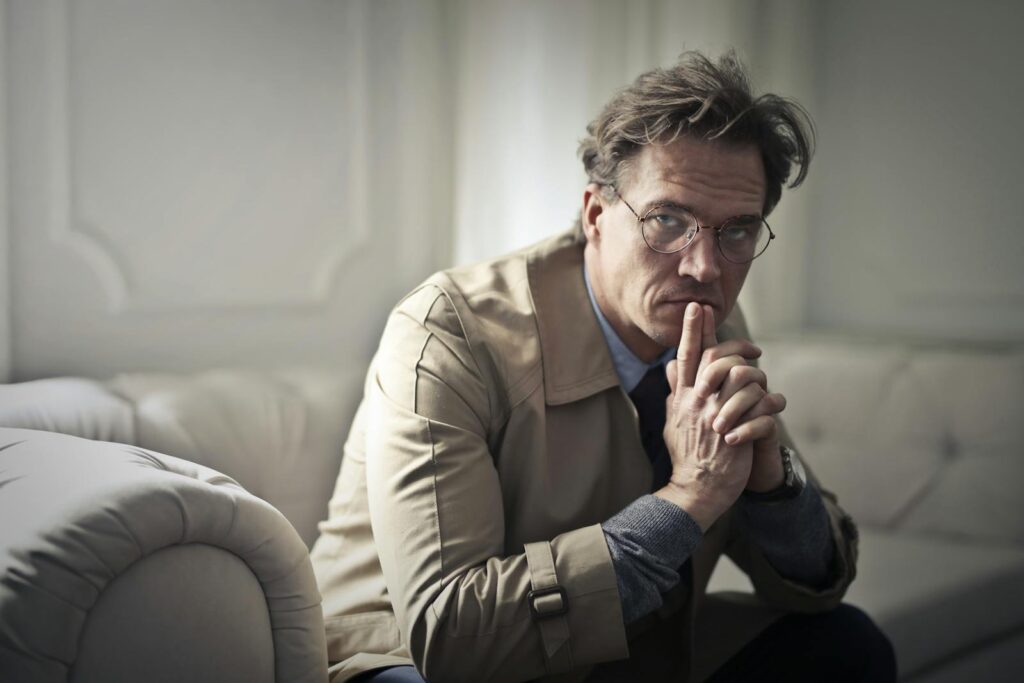 A thoughtful man in glasses sits pensively on a sofa in a well-lit room.