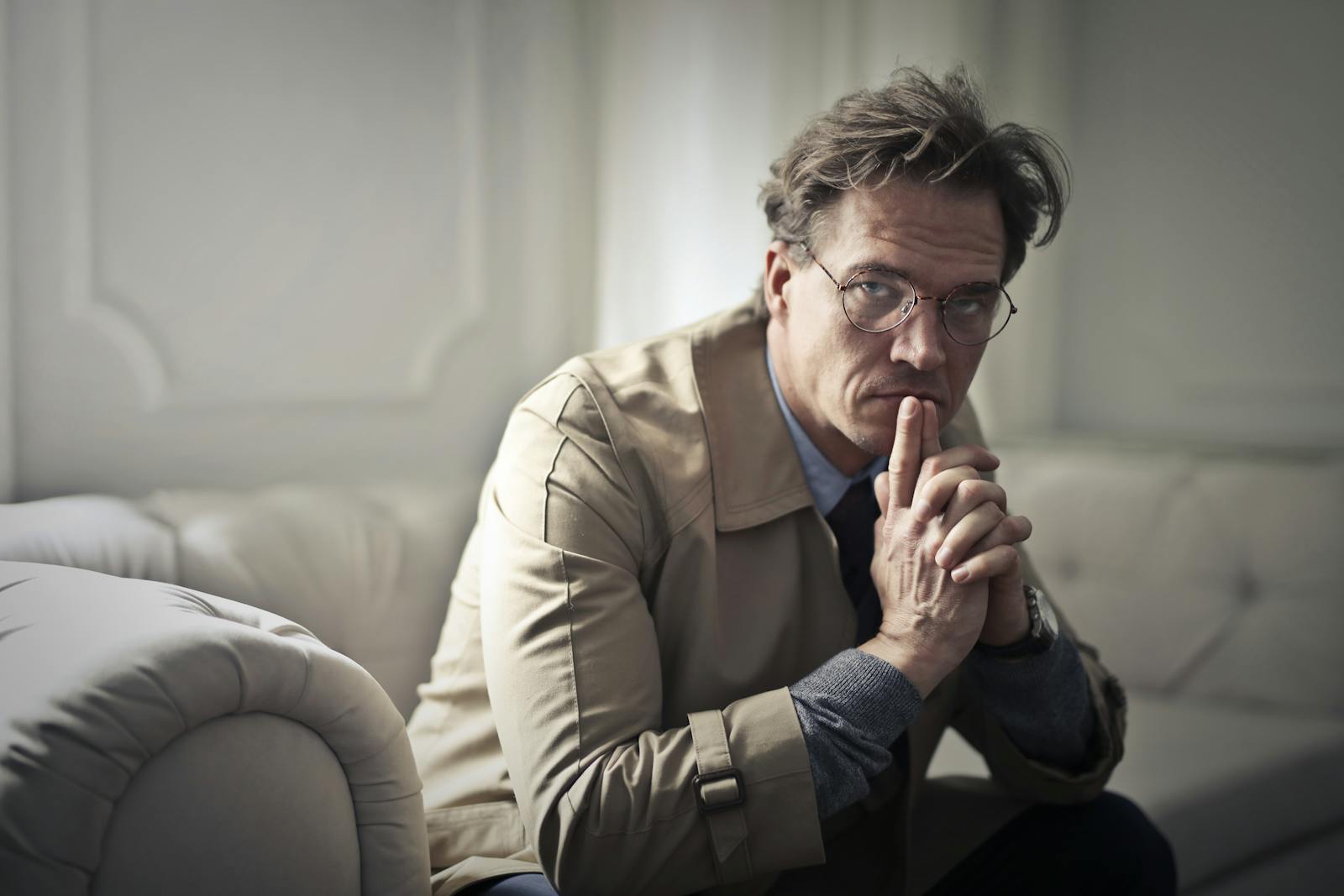 A thoughtful man in glasses sits pensively on a sofa in a well-lit room.