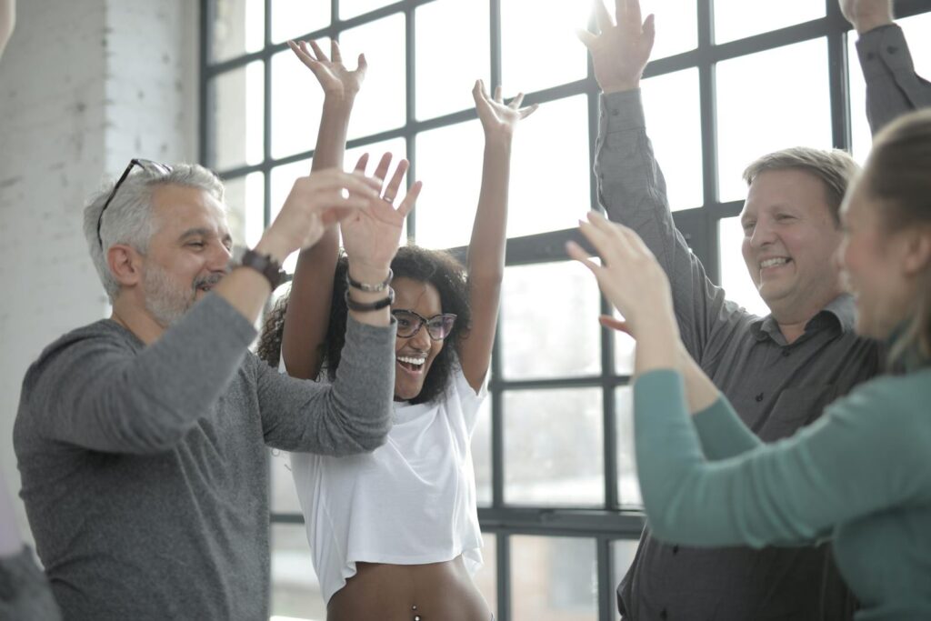 Diverse group of adults joyfully raising hands indoors, celebrating teamwork and camaraderie.