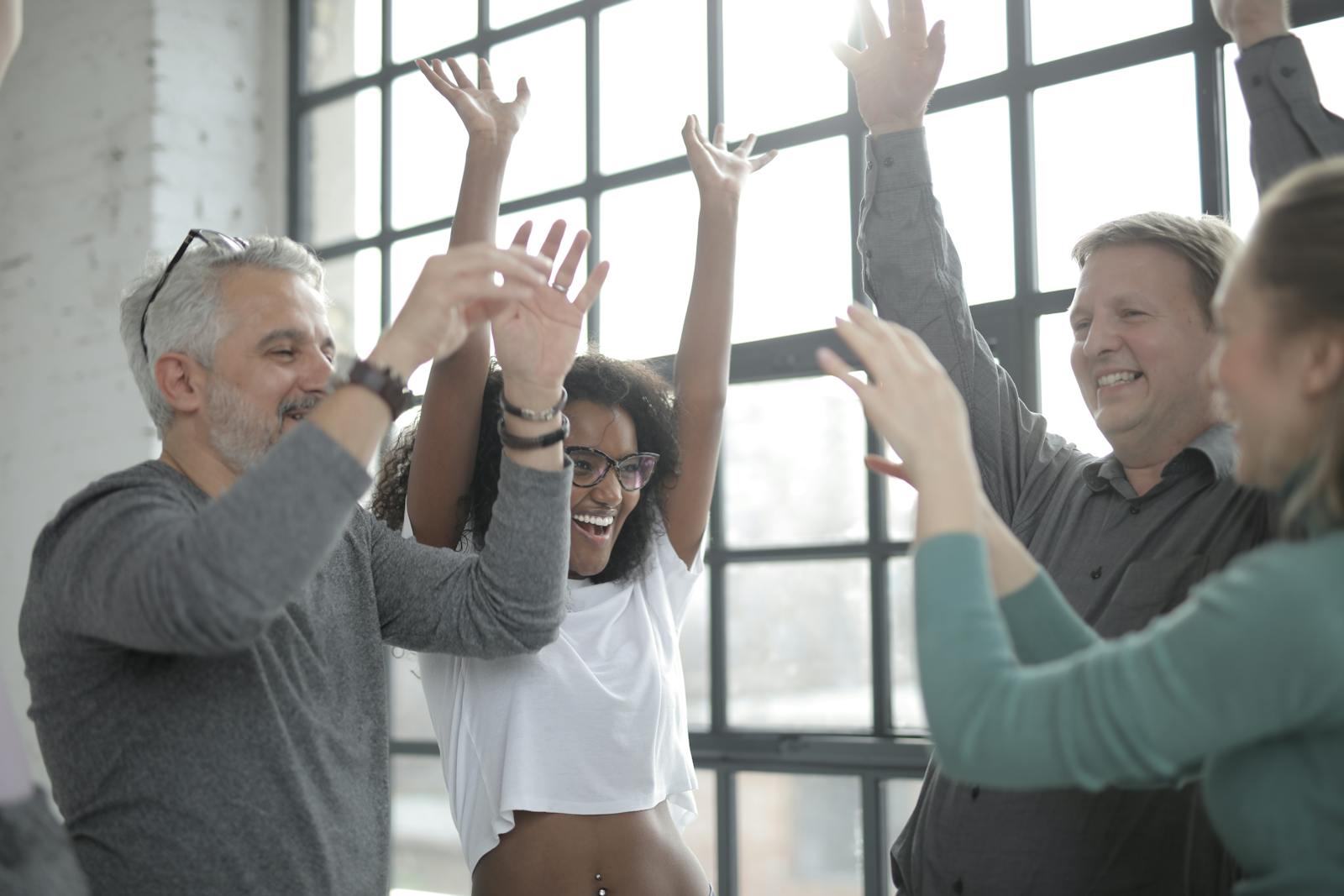 Diverse group of adults joyfully raising hands indoors, celebrating teamwork and camaraderie.