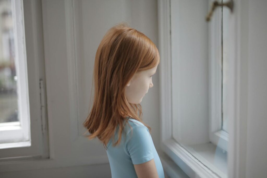 A young redhead girl thoughtfully gazing out a window inside a bright room.