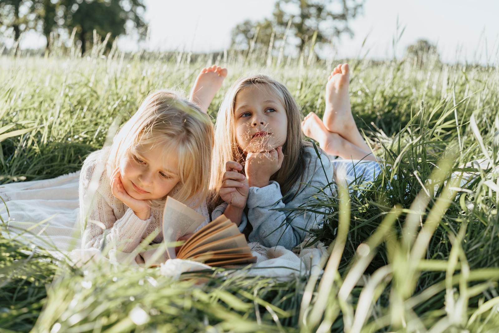 Two young girls lying on a picnic blanket in a sunny field, reading and daydreaming.