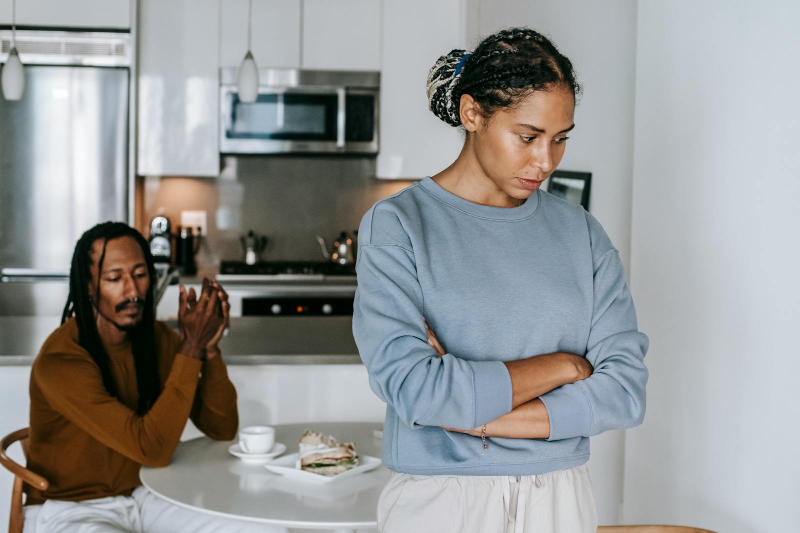 Young discontented African American female with folded arms against male partner at kitchen table during quarrel
