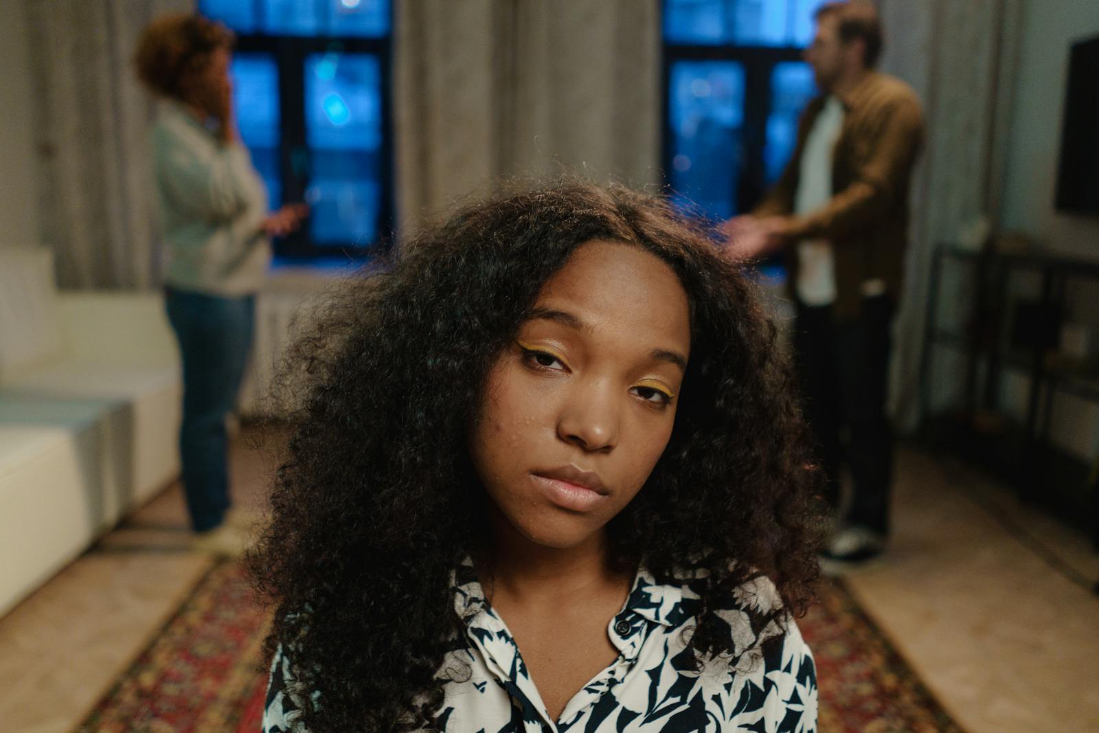 A teenage girl with curly hair looking concerned indoors with two adults in the background.