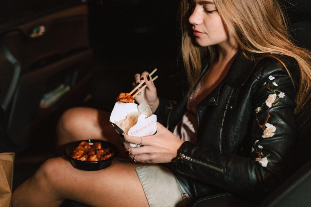 Young woman enjoying Asian takeout food while sitting in a car, wearing a black leather jacket.