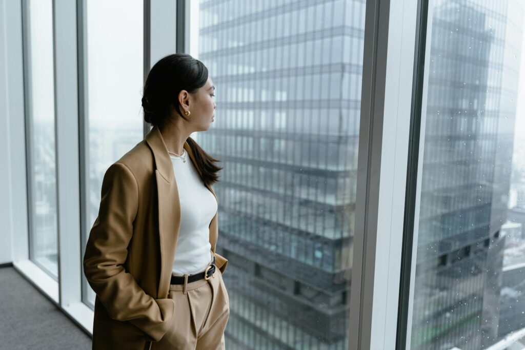 Businesswoman in brown coat looking out window in modern office.