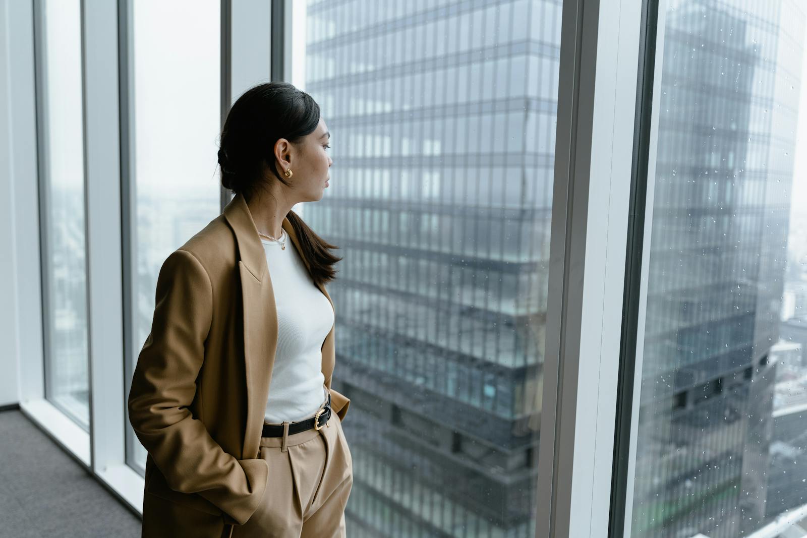 Businesswoman in brown coat looking out window in modern office.