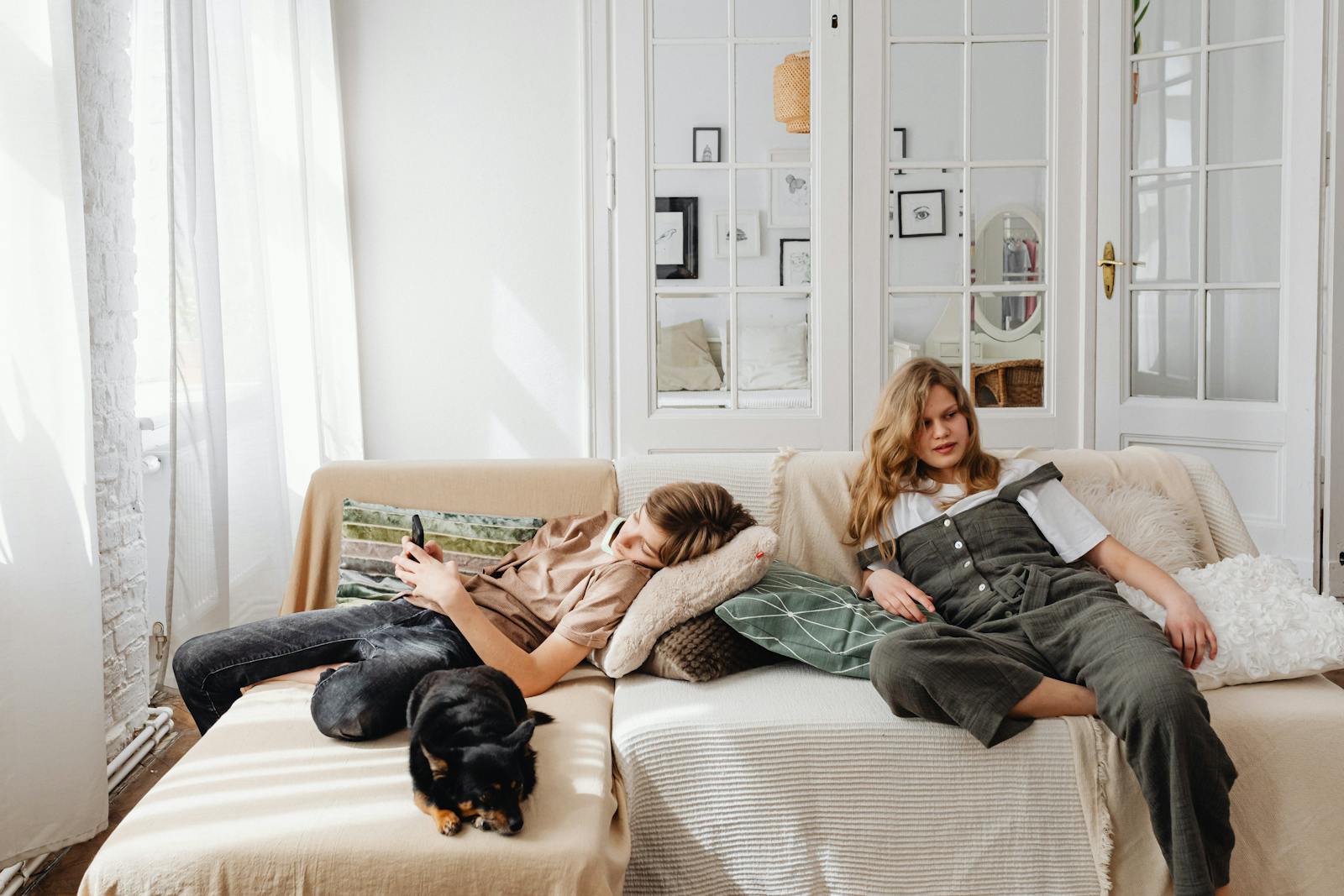 A brother and sister relaxing on a sofa with their black dog in a bright living room.