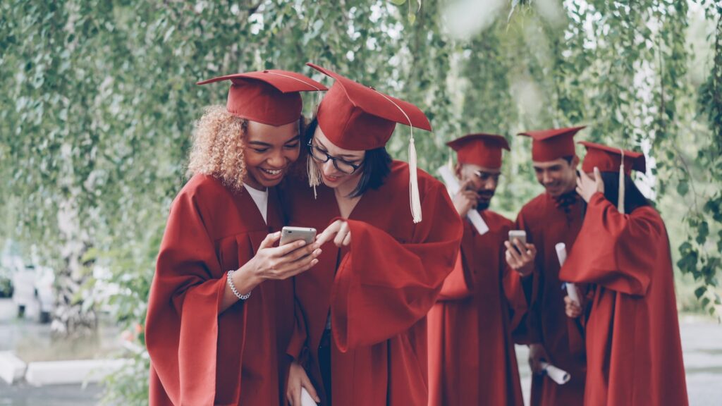 Graduates in red gowns look at a smartphone together.