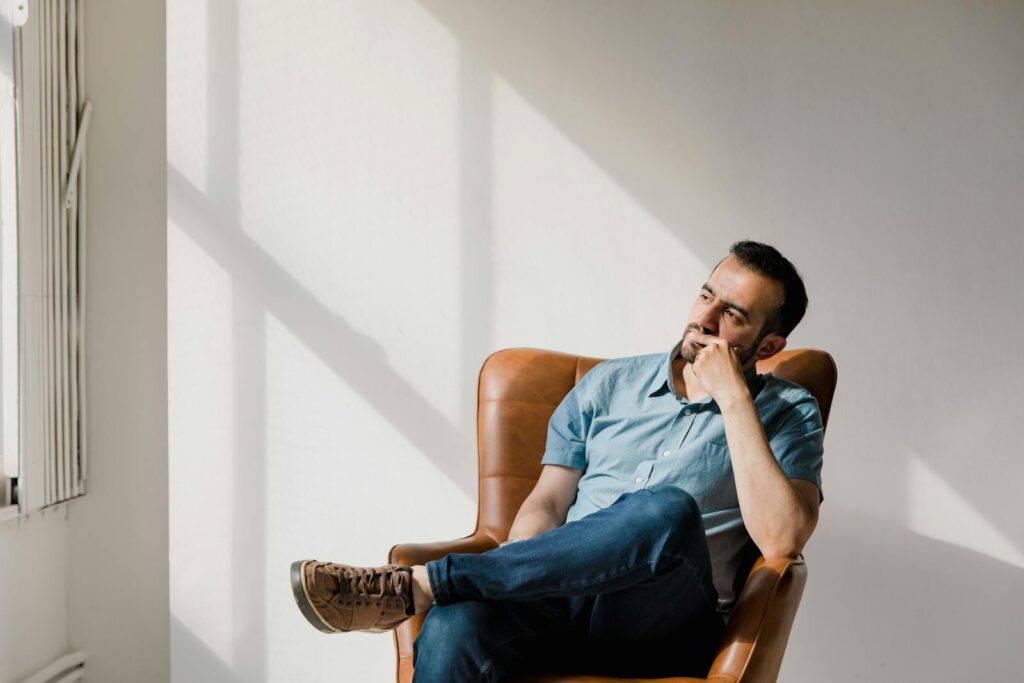 Man in casual attire sitting thoughtfully in a sunlit room, deep in contemplation.