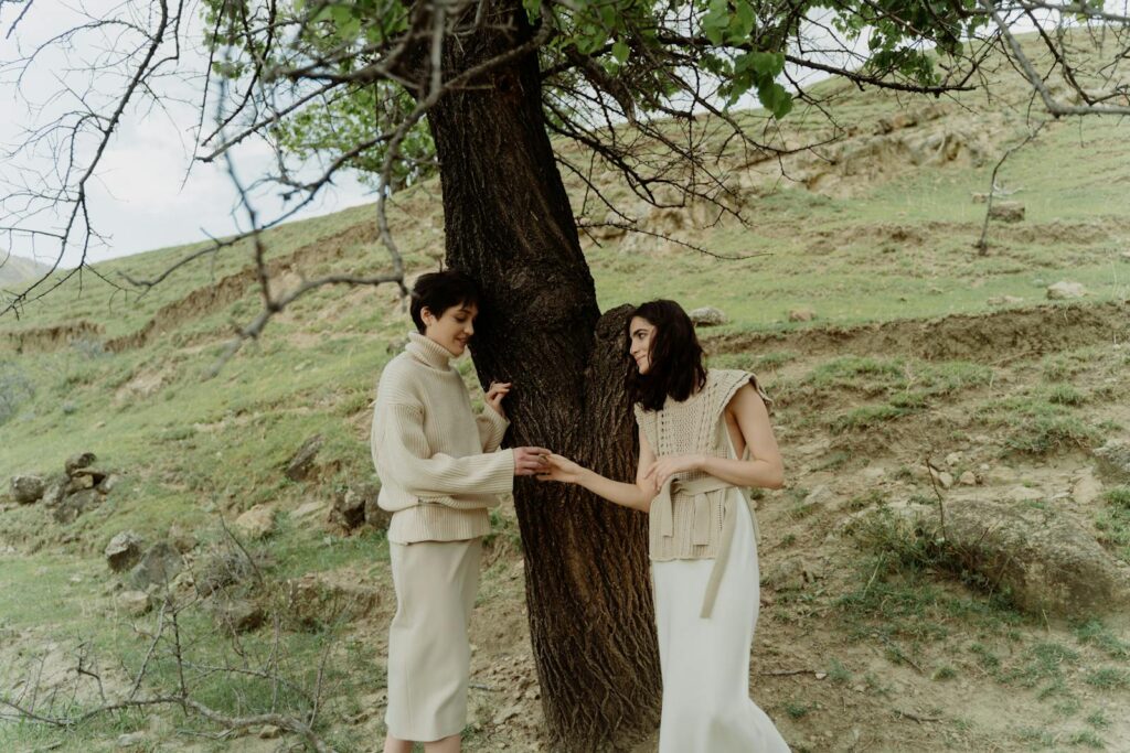 Lesbian couple holding hands under a tree, embracing love and nature in a serene outdoor setting.