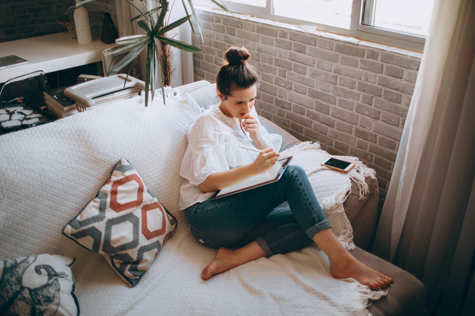 A woman sits on a cozy sofa by a window, writing in a notebook, reflecting a peaceful indoor lifestyle.