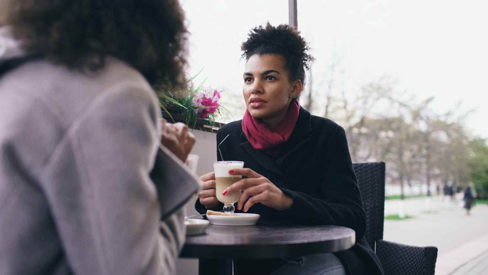 Two women having a conversation over coffee at an outdoor cafe on a bright day.