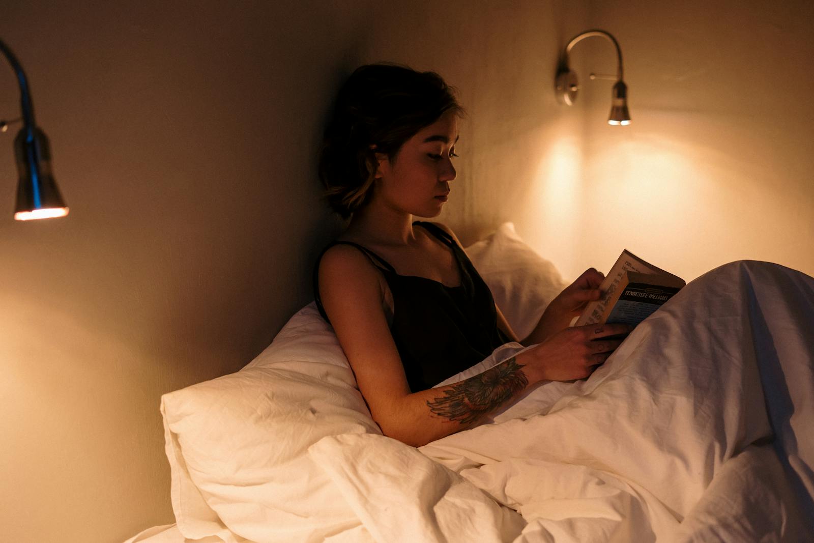 Woman with tattoo reading a book in bed under warm lamp light, creating a cozy atmosphere.