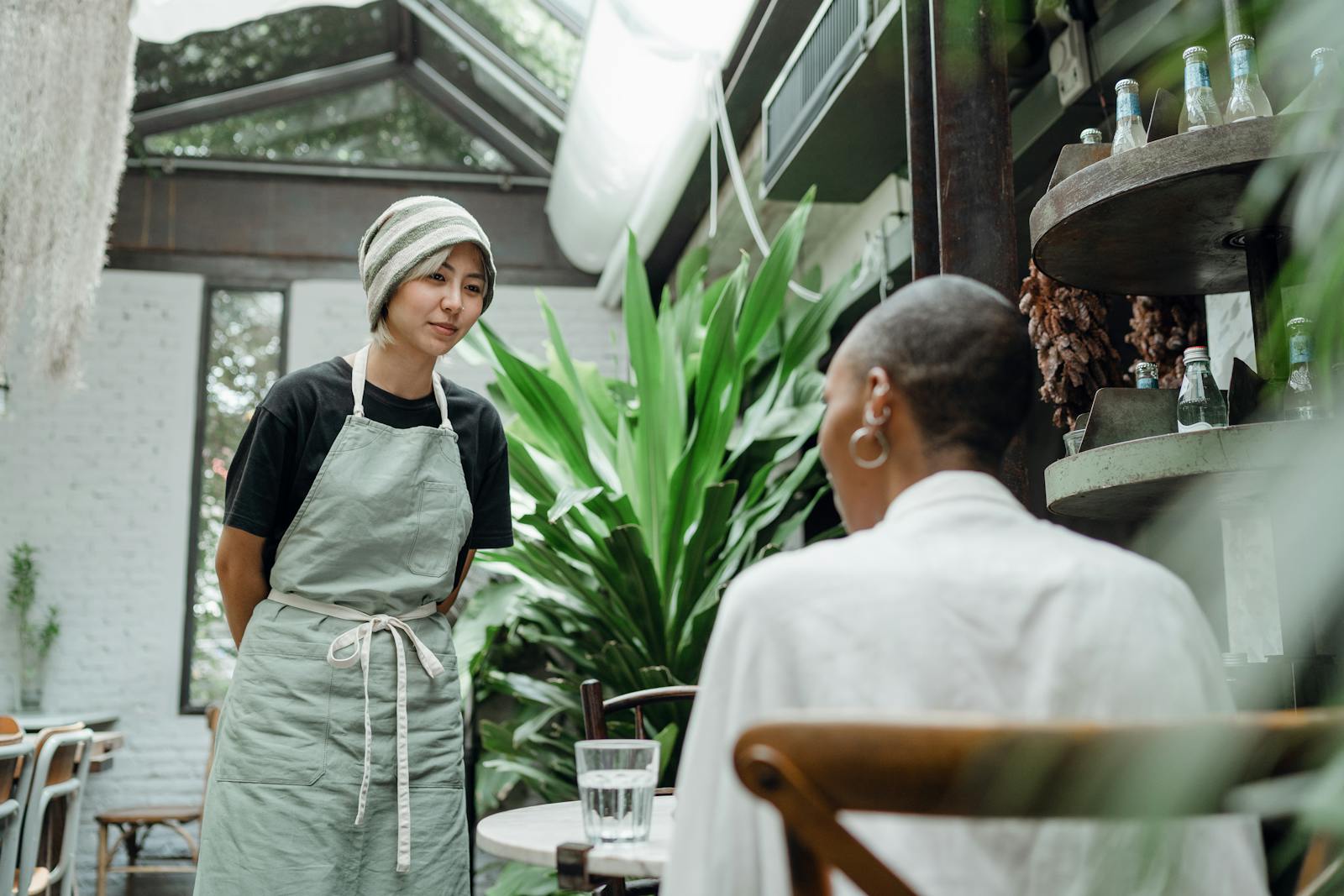 Friendly young Asian waitress in apron and hat standing near table and talking to female customer while working in cozy cafe