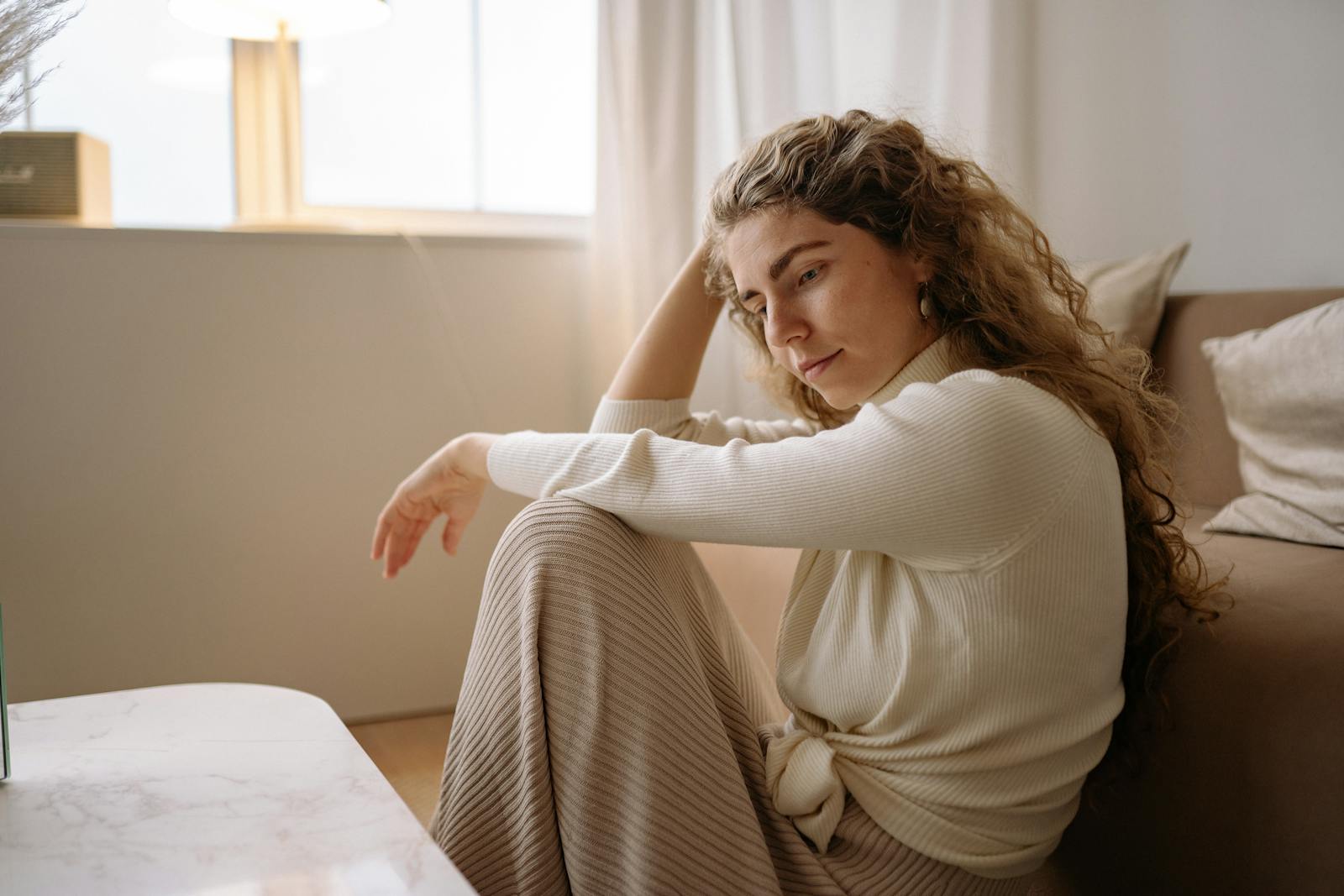 A woman in a cozy living room, sitting pensively with natural light streaming in. Calm and introspective atmosphere.