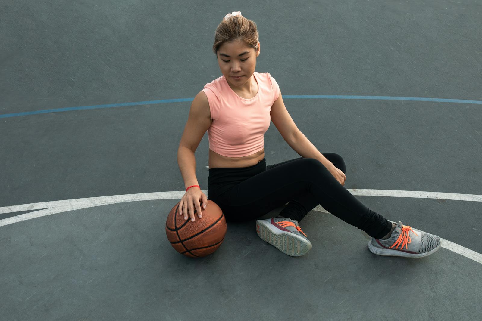 East Asian woman in sportswear sitting on an outdoor basketball court, holding a basketball.