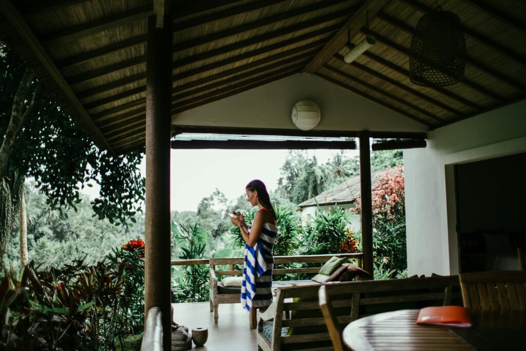 Woman wrapped in towel using phone on a tropical porch with lush greenery.