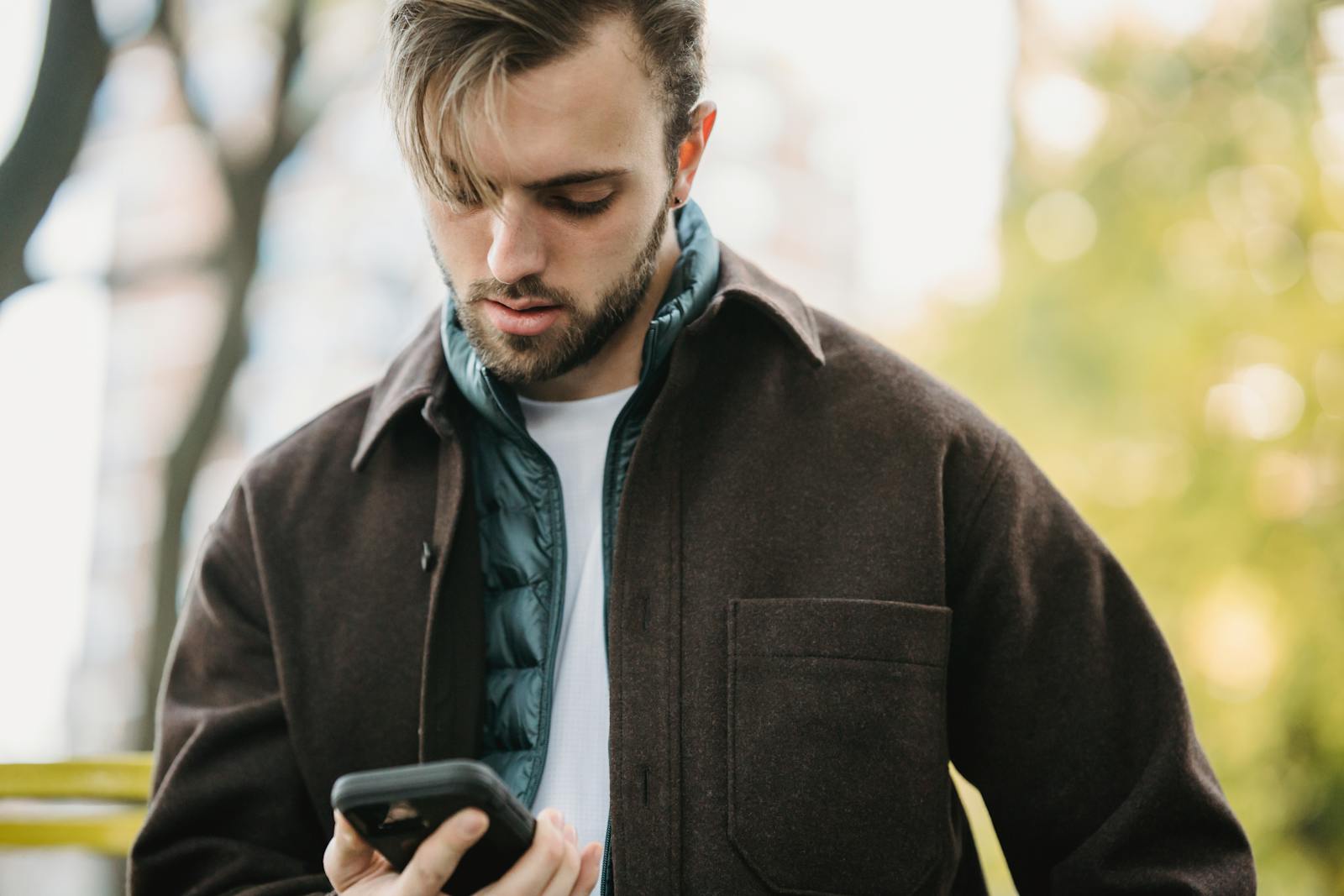 Young bearded man in casual clothes receiving text message on smartphone while standing on street