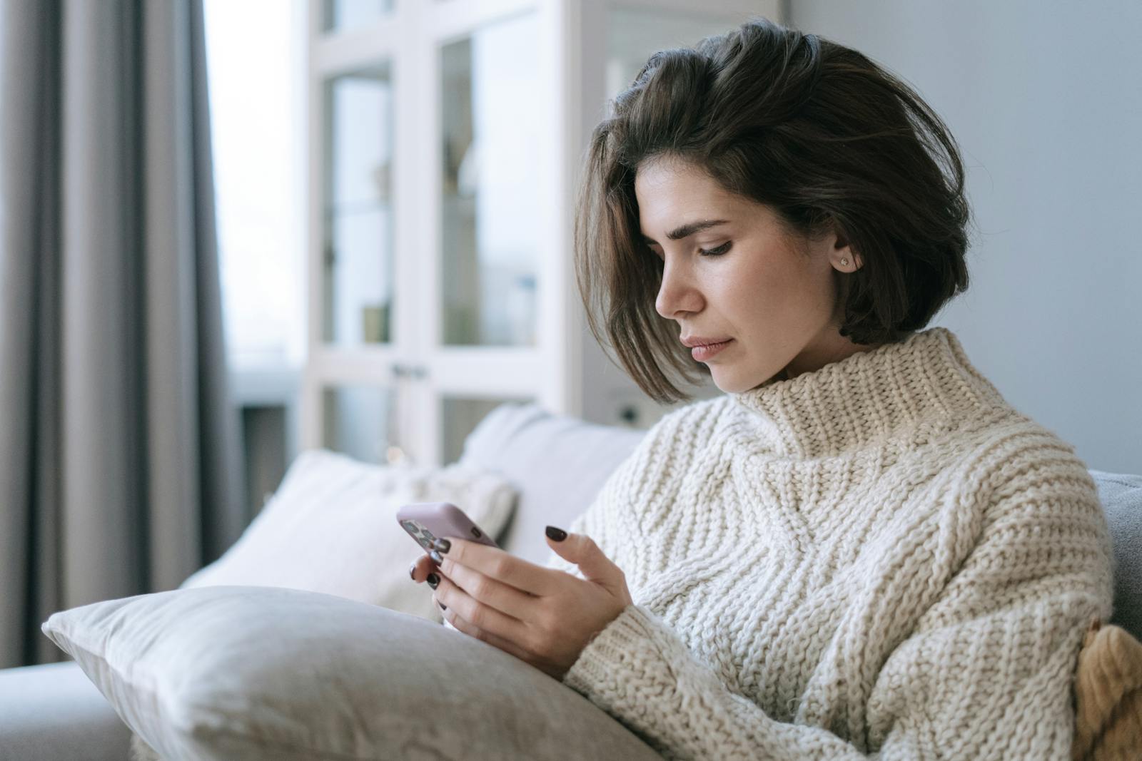 Woman in knitted sweater using smartphone on a cozy sofa indoors.