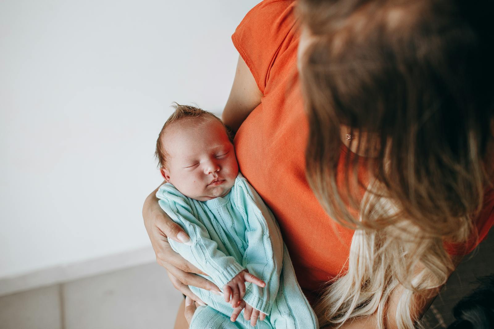 Crop anonymous mother with long hair with sleeping baby in arms sitting in light room near white wall at home
