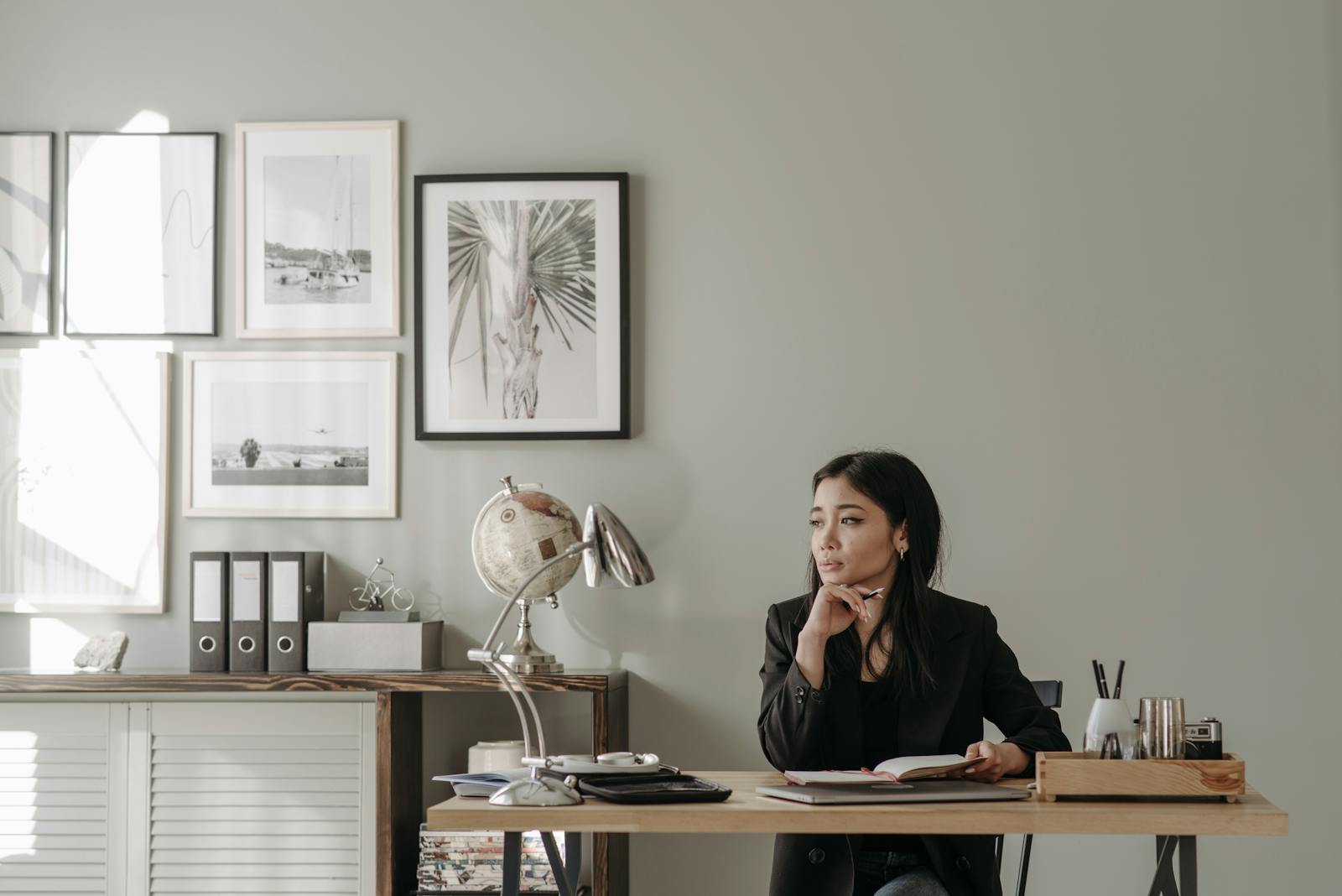 Asian woman in black blazer sitting at a desk in stylish home office, deep in thought.