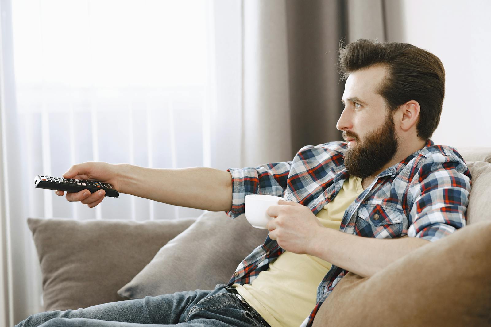 Bearded man in casual wear relaxing on a sofa, enjoying coffee while watching TV indoors.