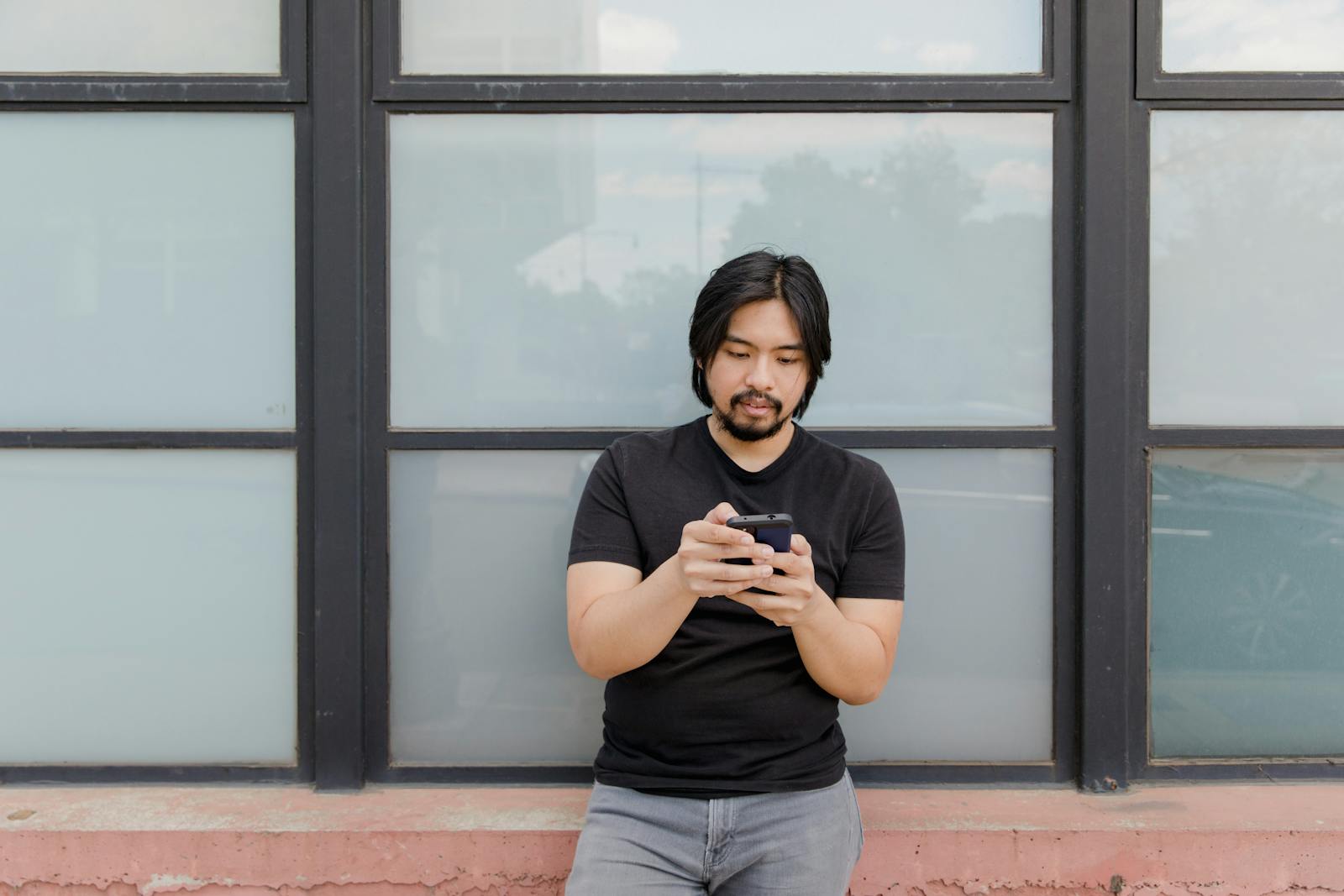 Asian man standing outdoors using phone against glass window backdrop.