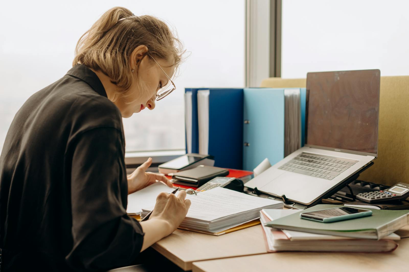 Woman in glasses intensely working at a desk with documents and a laptop.