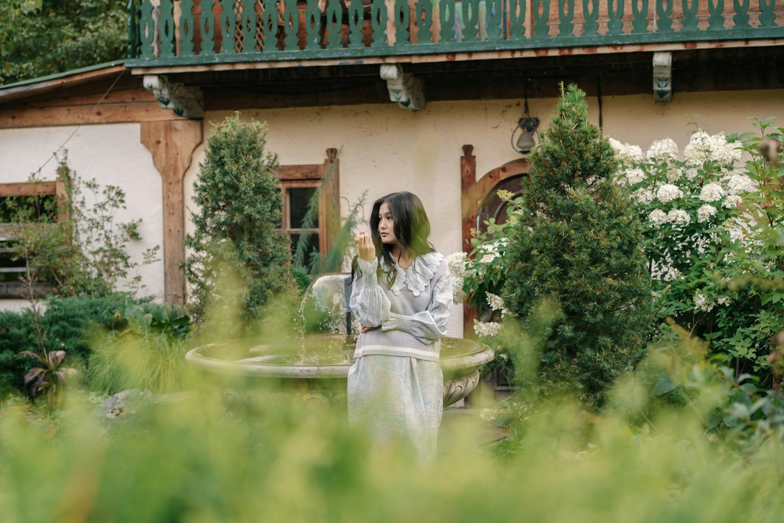 Asian woman in a vintage garden setting by a fountain, surrounded by greenery.