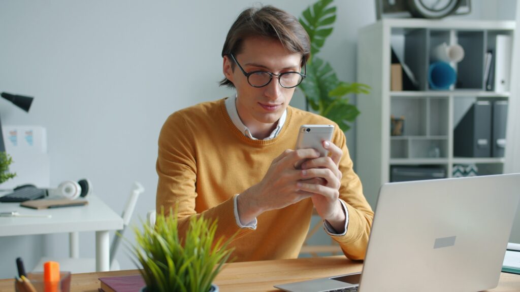 Man in glasses using phone at desk with laptop.