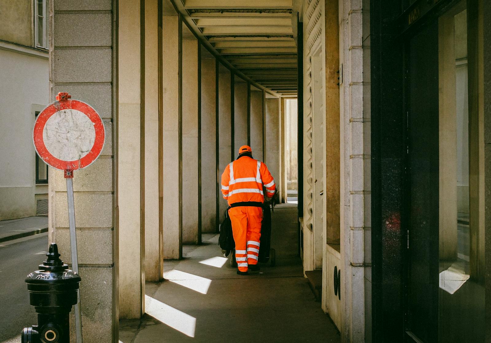 Street worker in orange uniform pushing a cart down a narrow urban alley during the day.