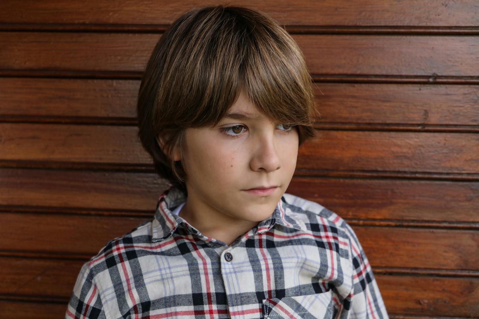 Portrait of a young boy in a plaid shirt against a wooden background, looking thoughtful.