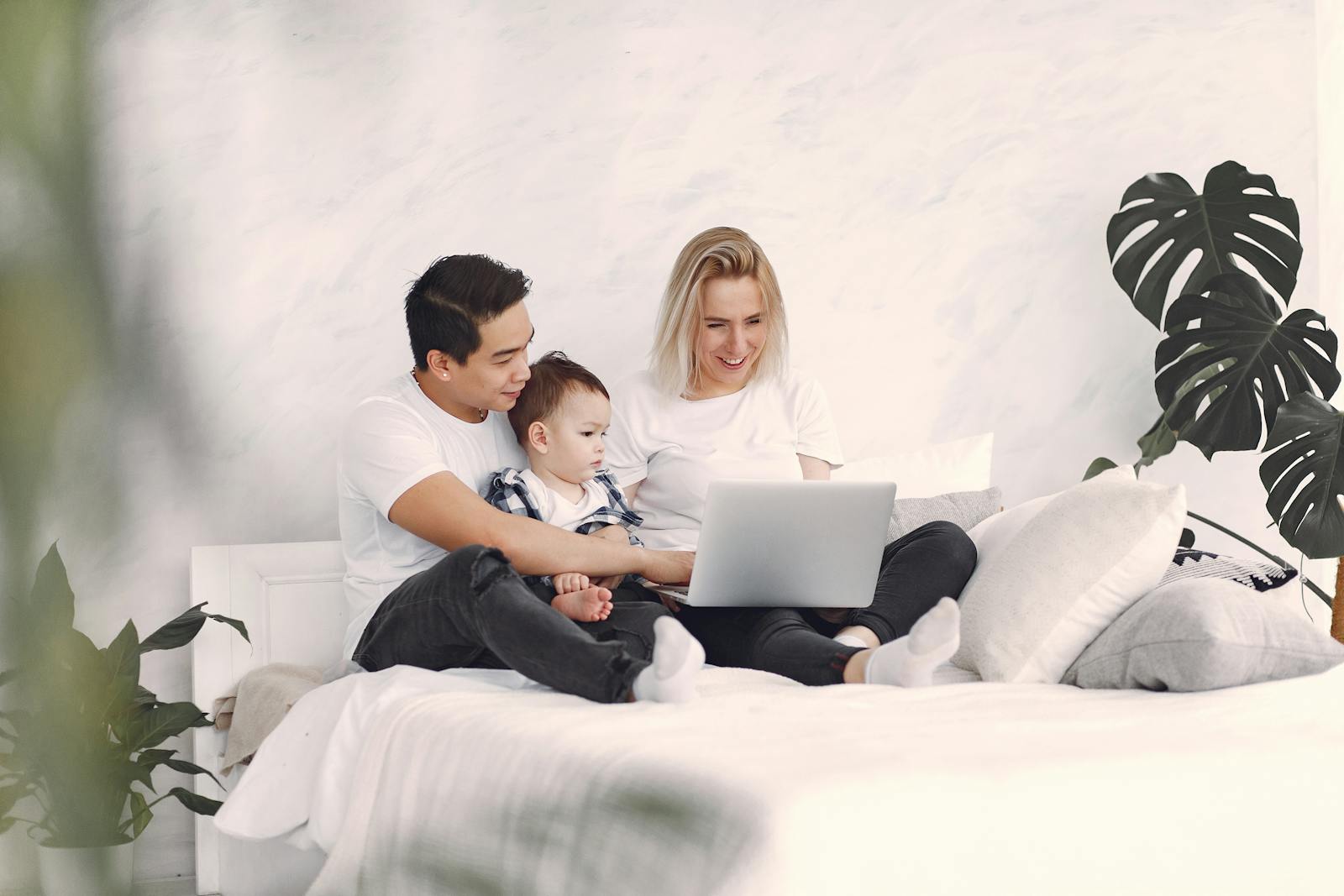 A happy family of three enjoying quality time on bed with a laptop, surrounded by plants.