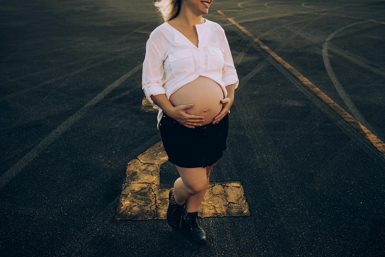 Expectant mother holding belly on a sunlit road, wearing casual attire.