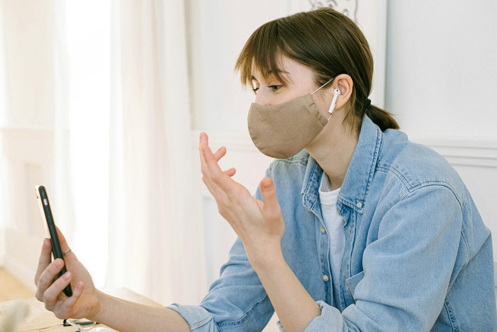 A woman wearing a face mask engages in a video call on her smartphone indoors.