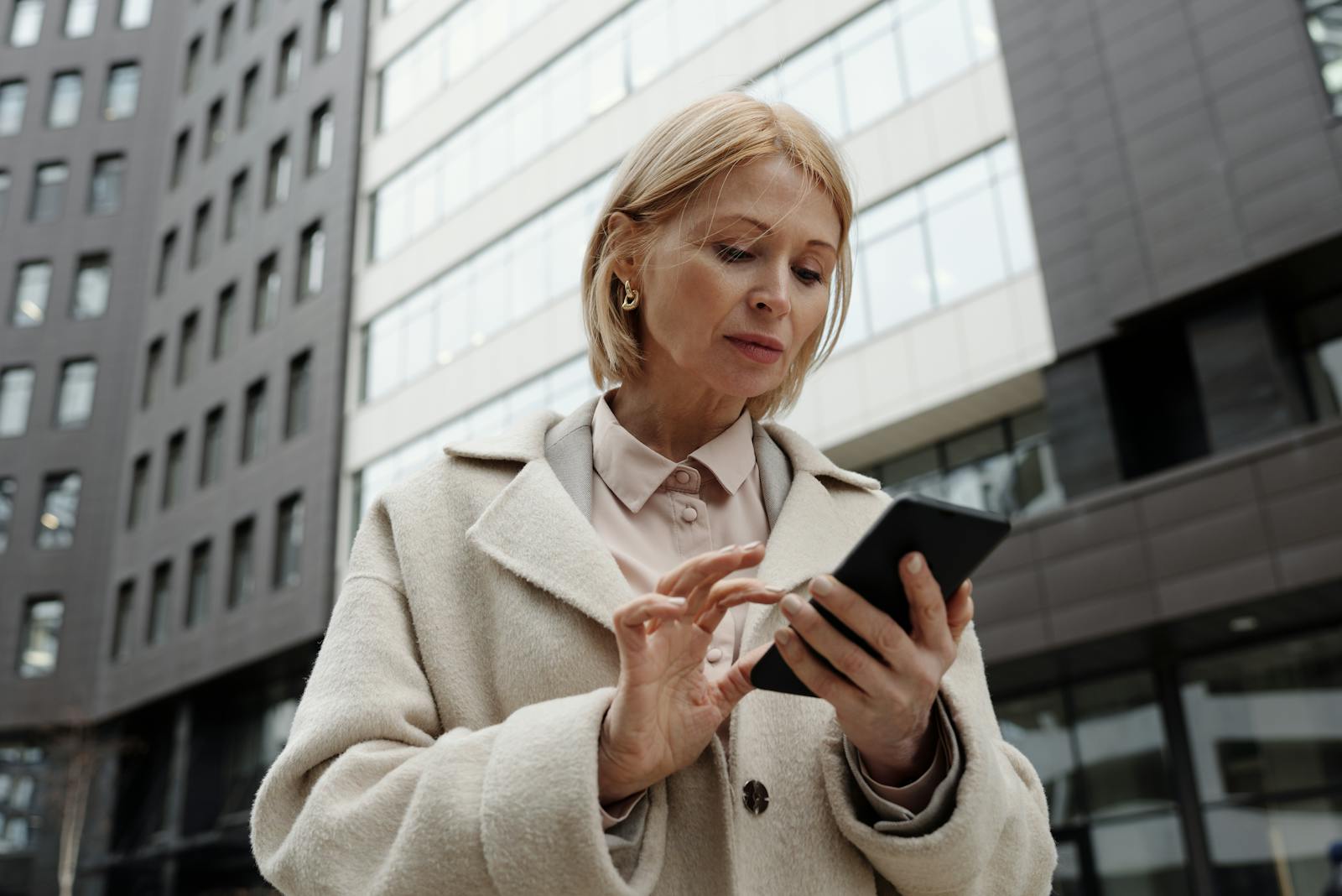 Woman in coat using smartphone in front of modern building. Professional and focused expression.