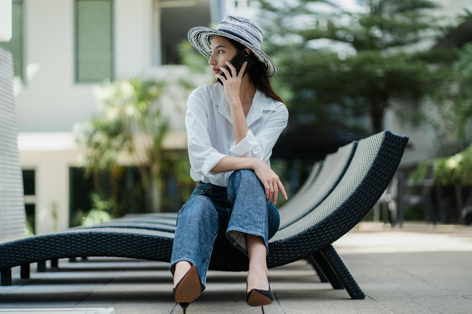 Full body of young stylish ethnic female in high heeled shoes and hat sitting on sunbed and having phone conversation in hotel garden