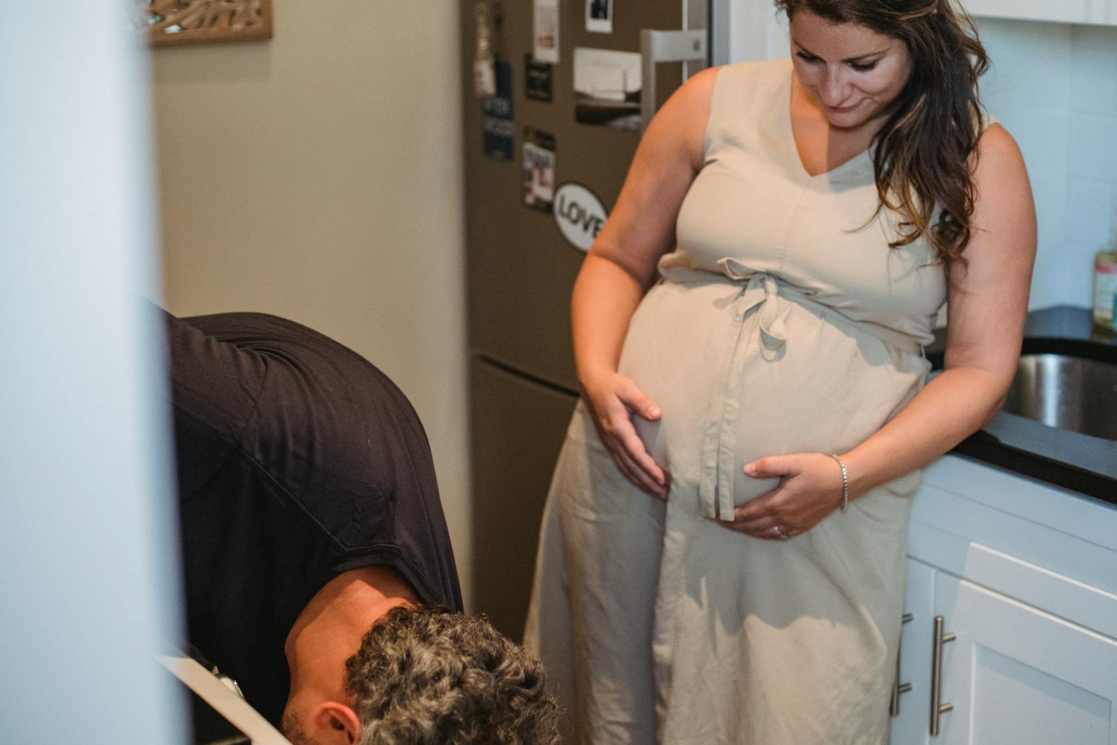 Happy adult woman in casual dress touching pregnant tummy while standing at kitchen and looking at husband leaning forward near oven