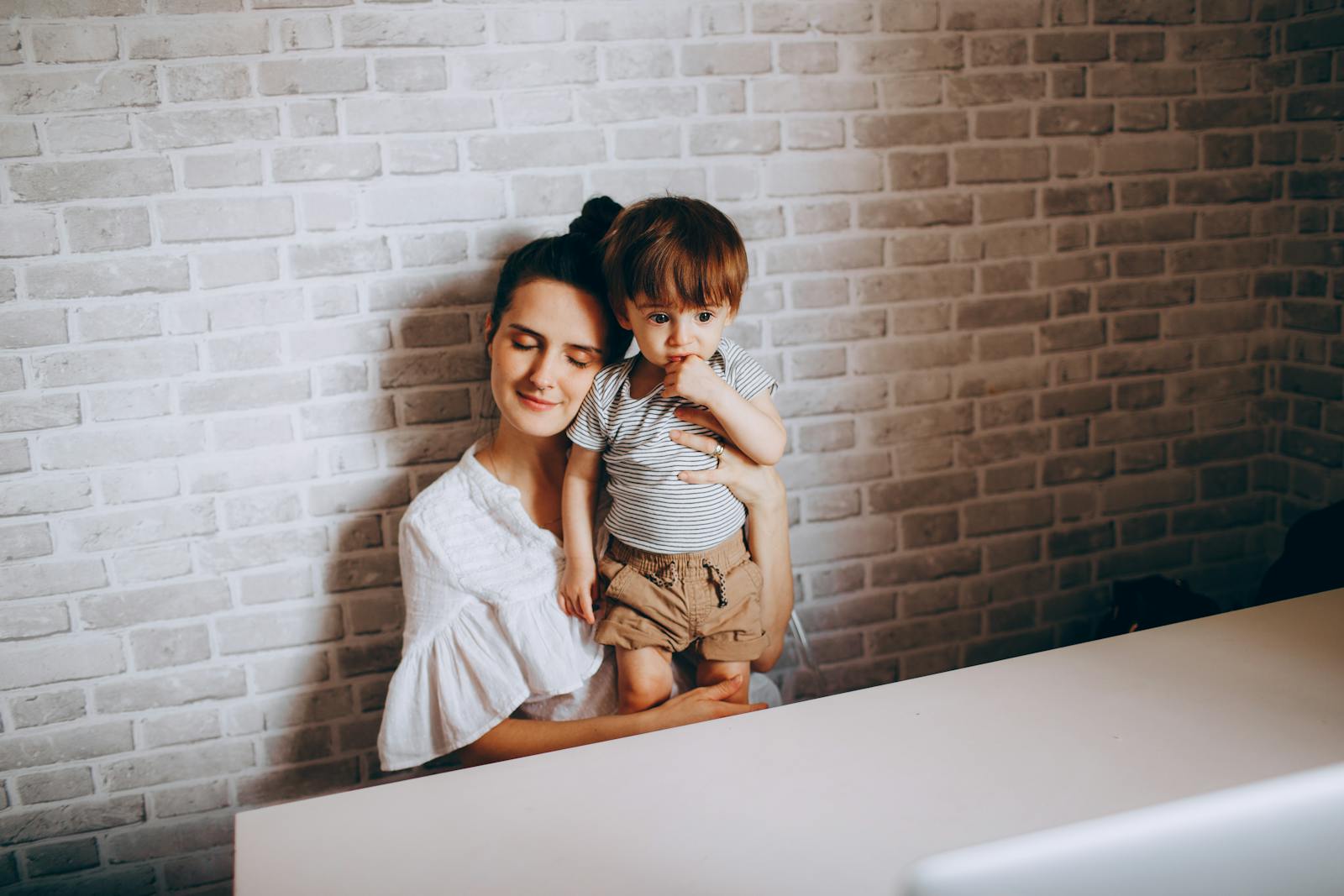Pleasant caring mother with eyes closed embracing adorable curious funny toddler near brick wall