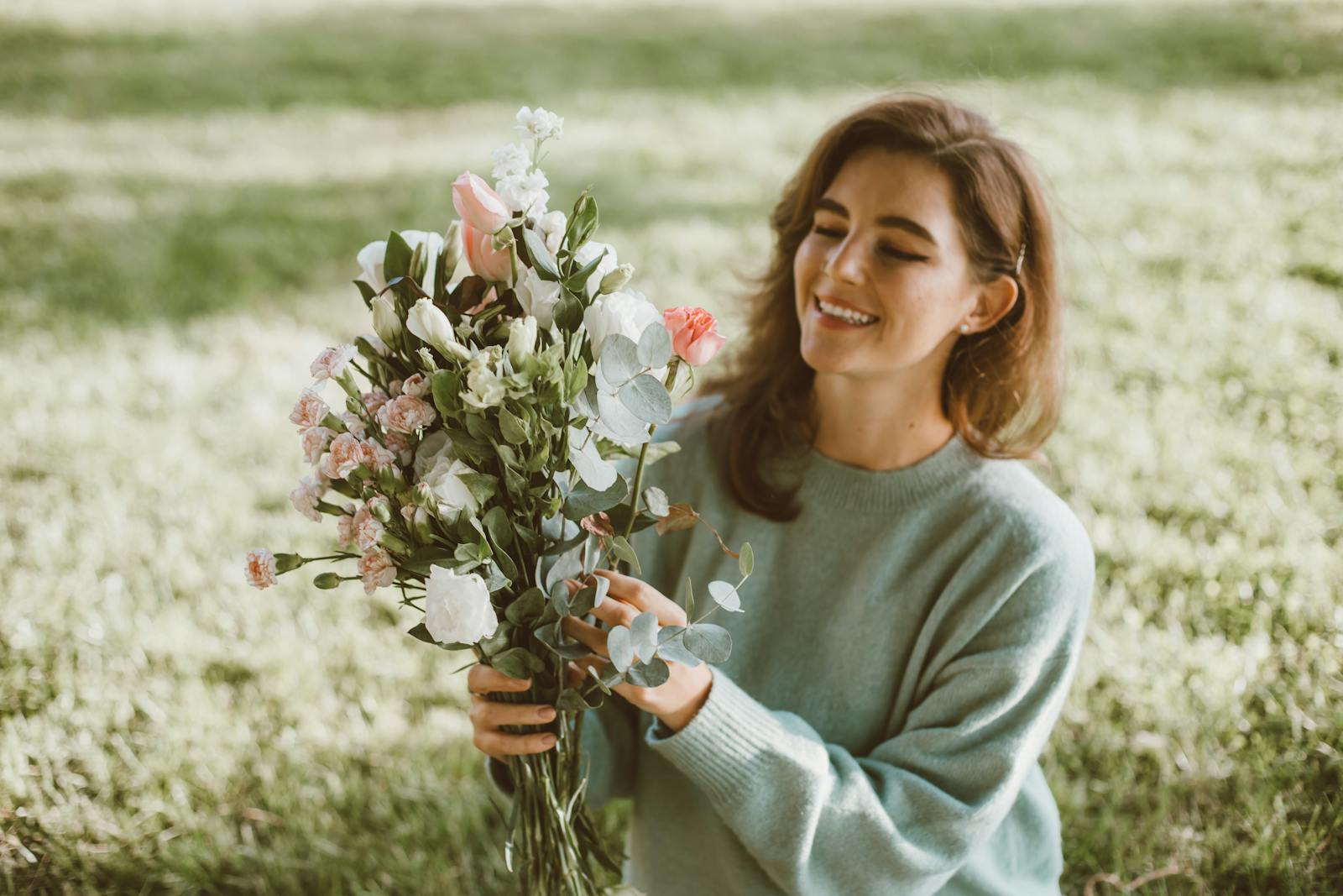 Smiling woman in a sweater holding a bouquet of mixed flowers in a sunny outdoor setting.