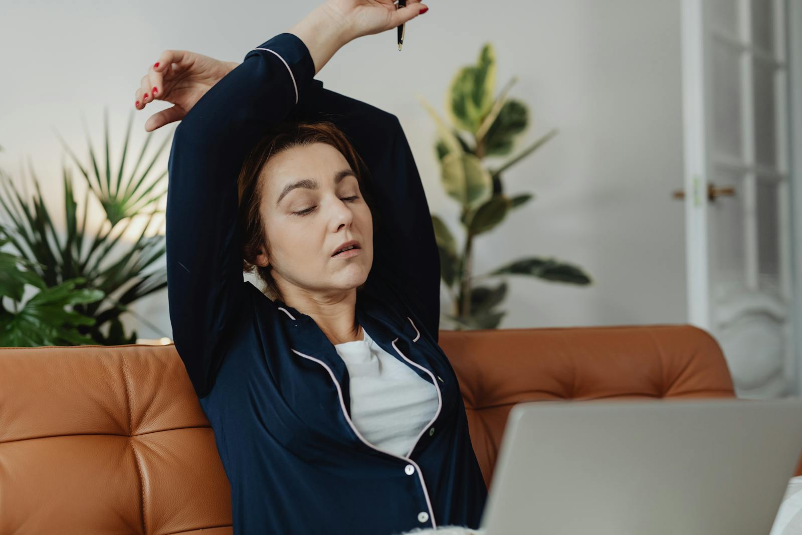 A tired woman in pajamas stretches while working remotely from her living room couch.