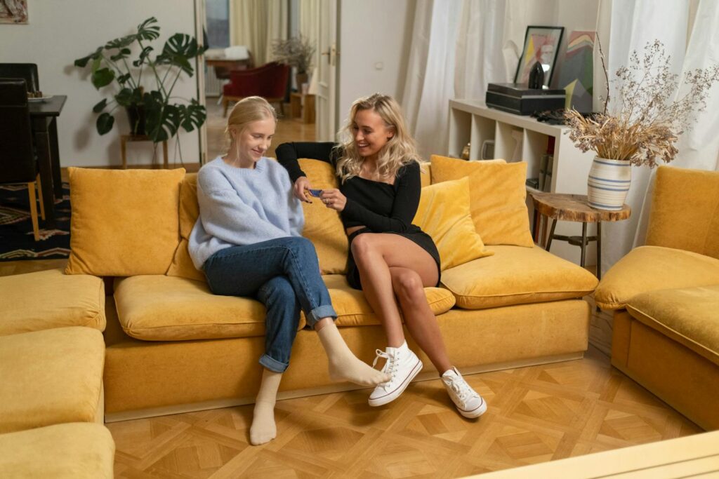 Two women sitting and smiling on a mustard yellow couch in a cozy living room.