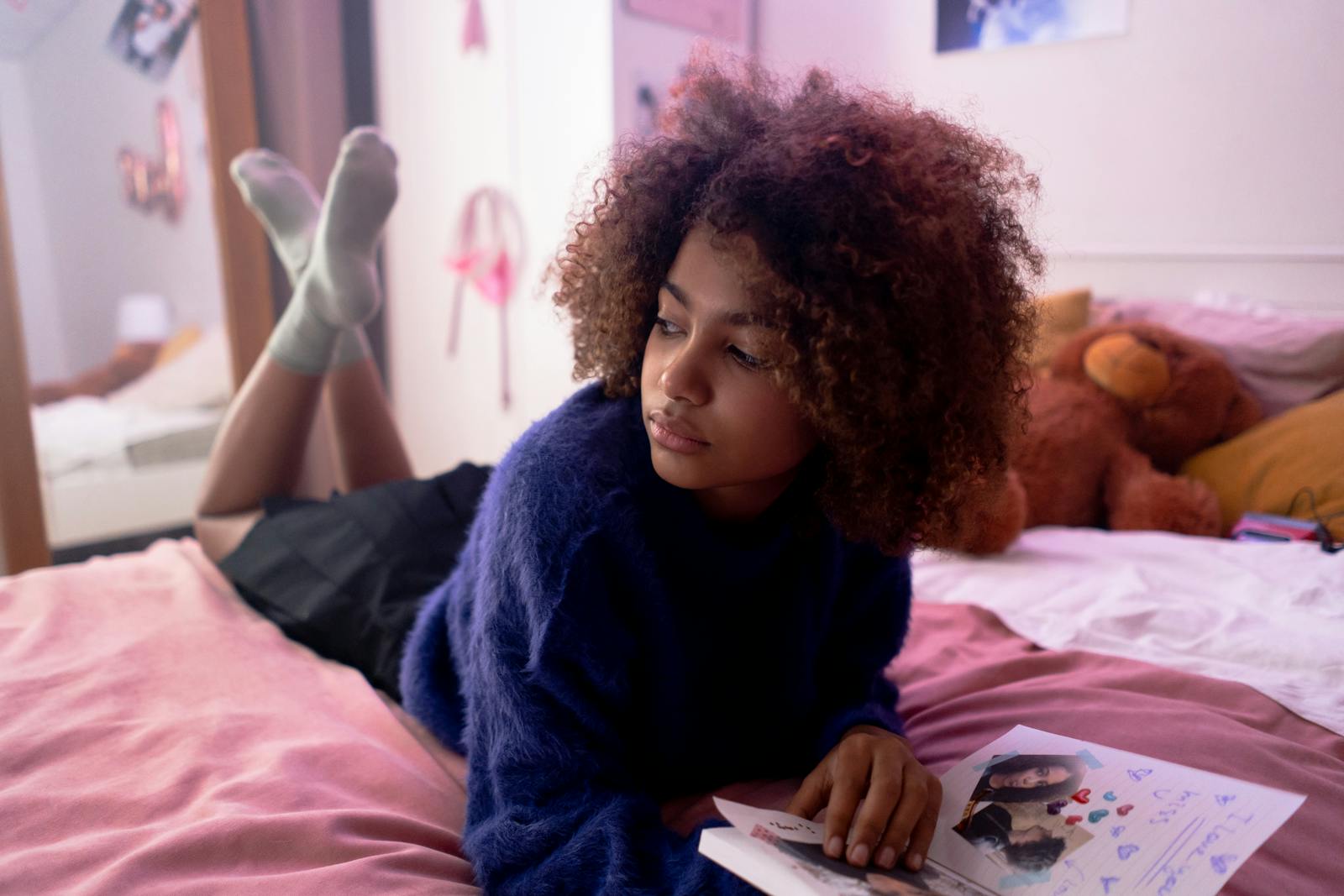 Teen girl with afro hair relaxing on bed with a notebook, surrounded by cozy decor and teddy bear.