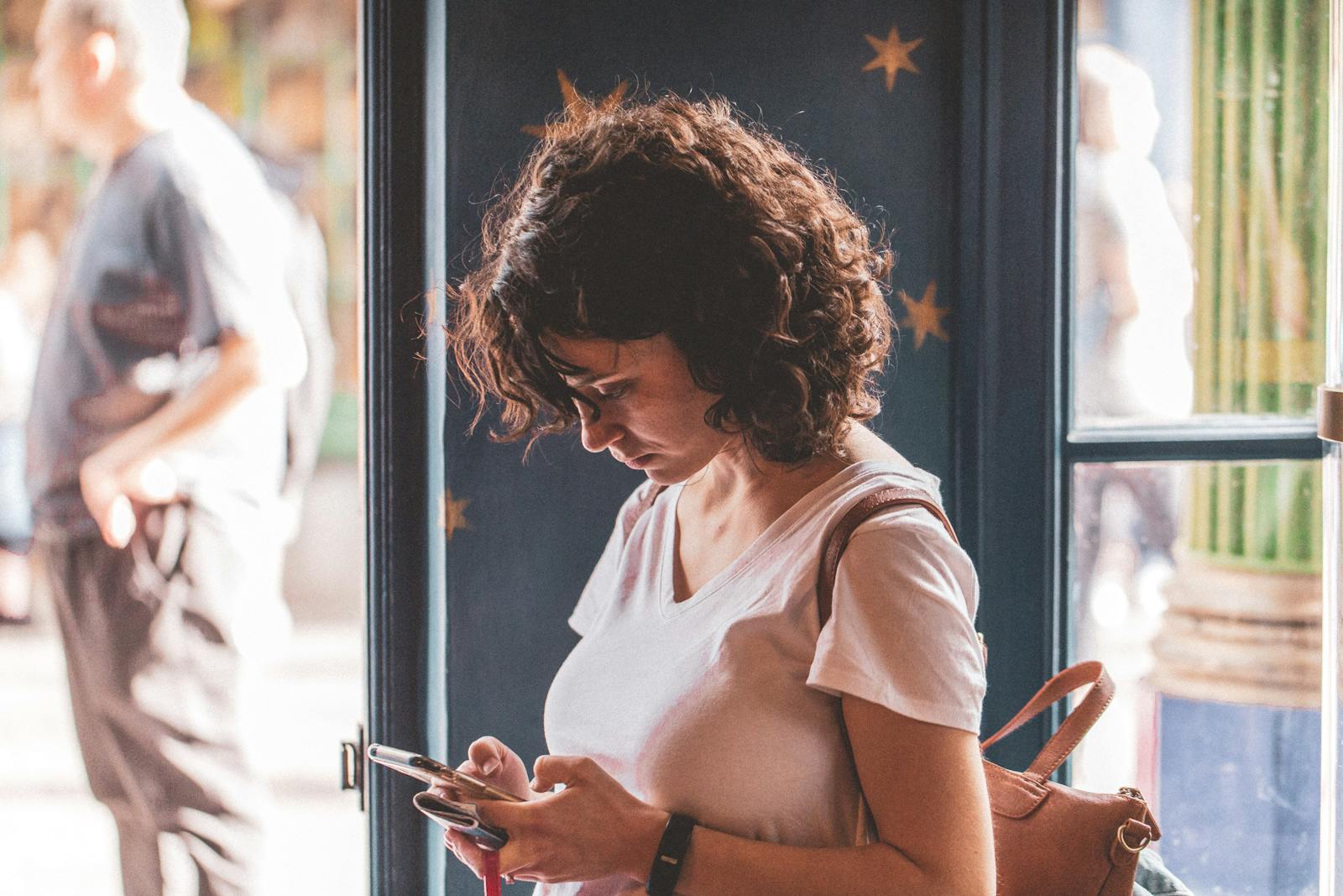 Woman with curly hair using smartphone by a sunlit window, wearing a white t-shirt.
