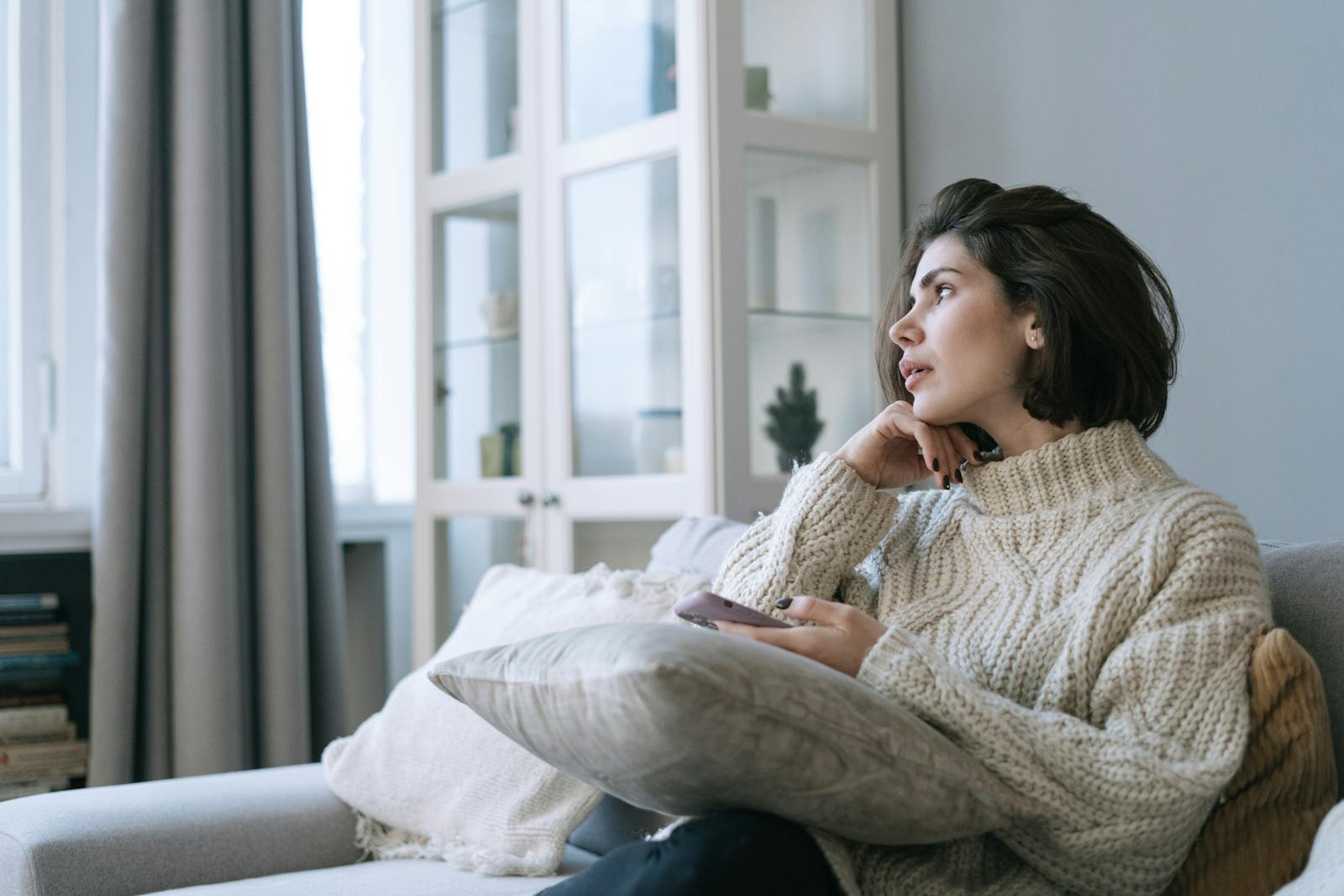 Woman in a comfy sweater sitting on couch, looking thoughtful with a smartphone in hand.