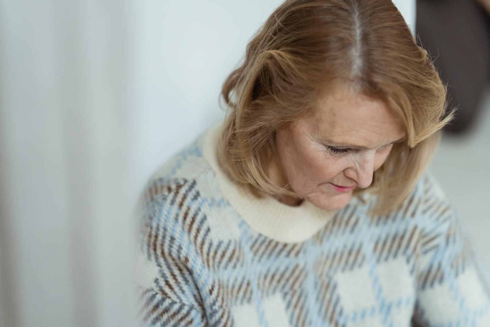 Crop positive middle aged lady with blond hair in stylish sweater smiling and looking down in daylight