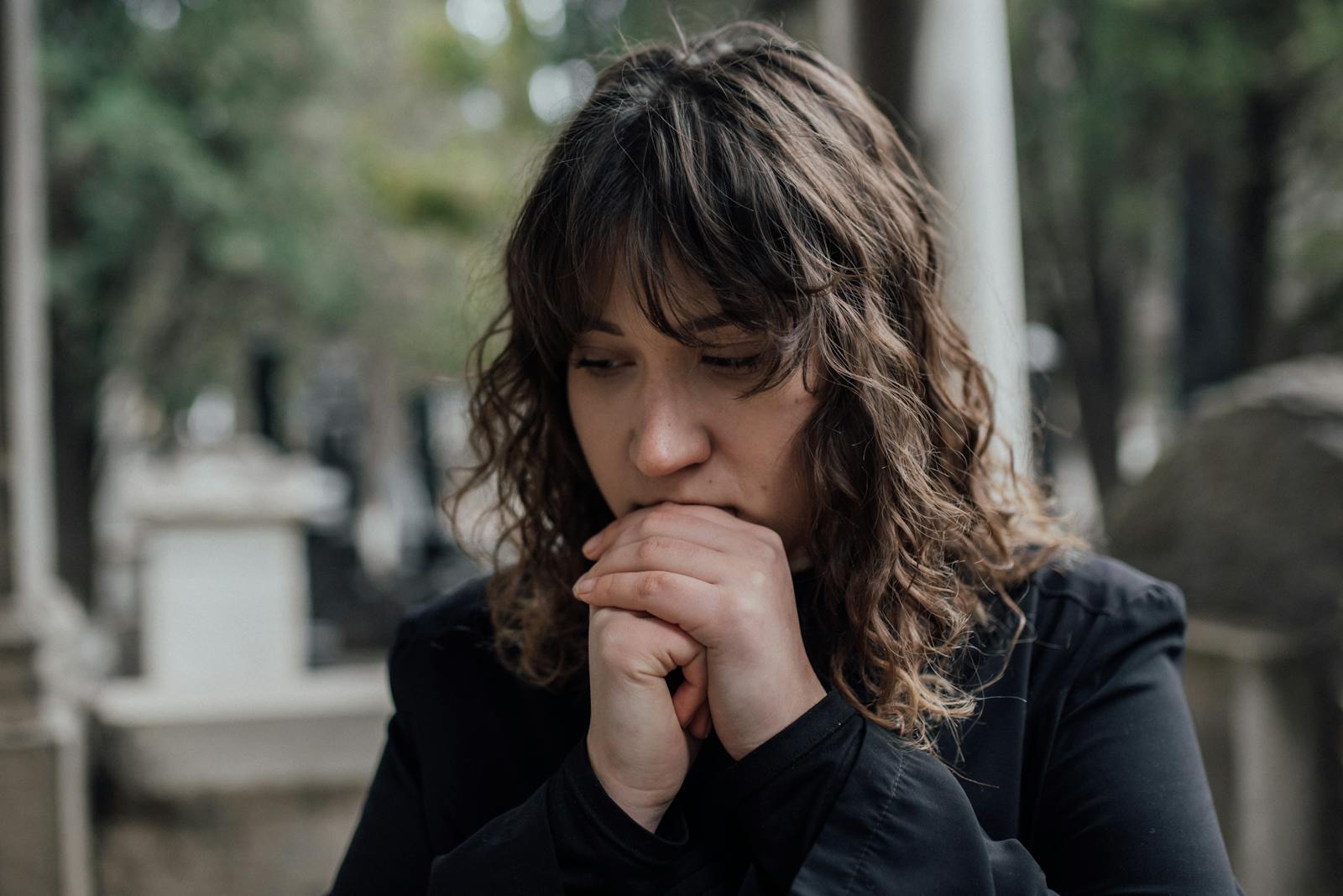 Pensive woman in black coat standing outdoors with hands together, conveying sadness.