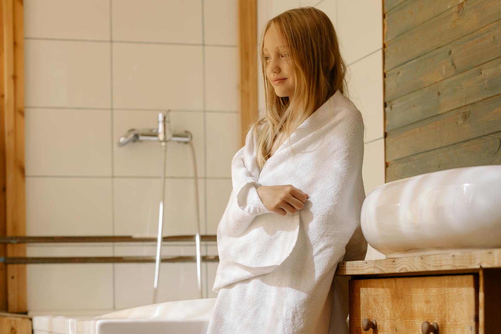 A young blonde girl wrapped in a towel standing by a bathroom sink, in a warm and cozy setting.